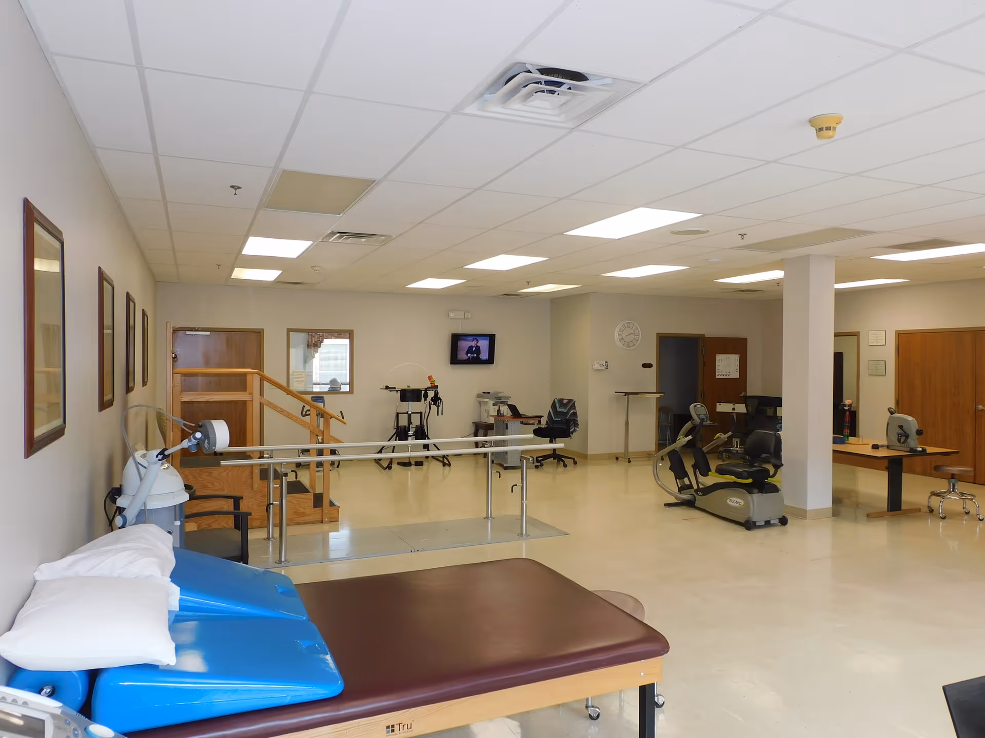 Spacious rehabilitation therapy room with treatment tables, parallel bars, and exercise equipment.