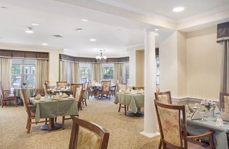 Spacious dining room with multiple tables covered in green tablecloths, set with napkins and floral centerpieces under soft overhead lighting.