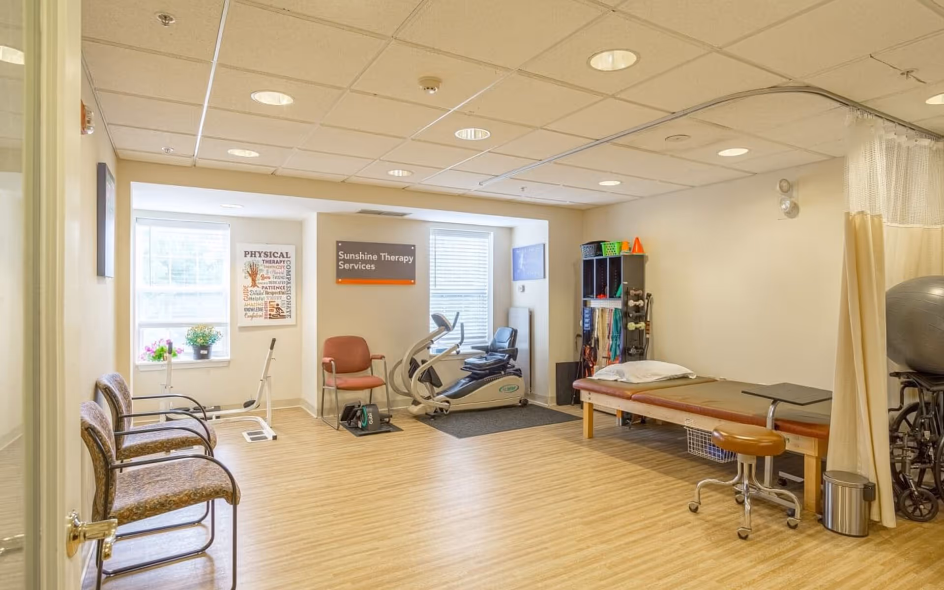 Physical therapy room with exercise equipment, a treatment table, chairs and shelving under bright windows.