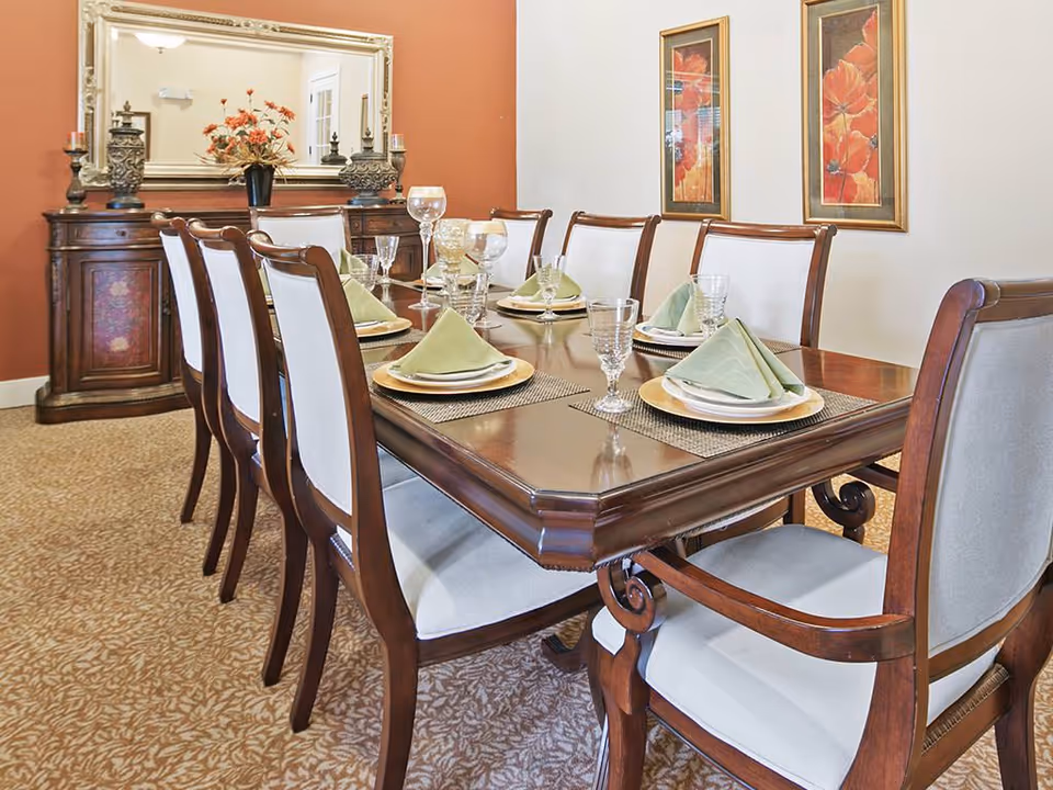 A formal dining room with a long wooden table set for eight people. Each place setting includes a gold charger plate, white plates, a folded green napkin, and a clear drinking glass. The chairs have wooden frames with light-colored upholstered seats and backs. In the background, there is a wooden sideboard with decorative items and a large mirror above it. Two framed floral paintings hang on the wall to the right.