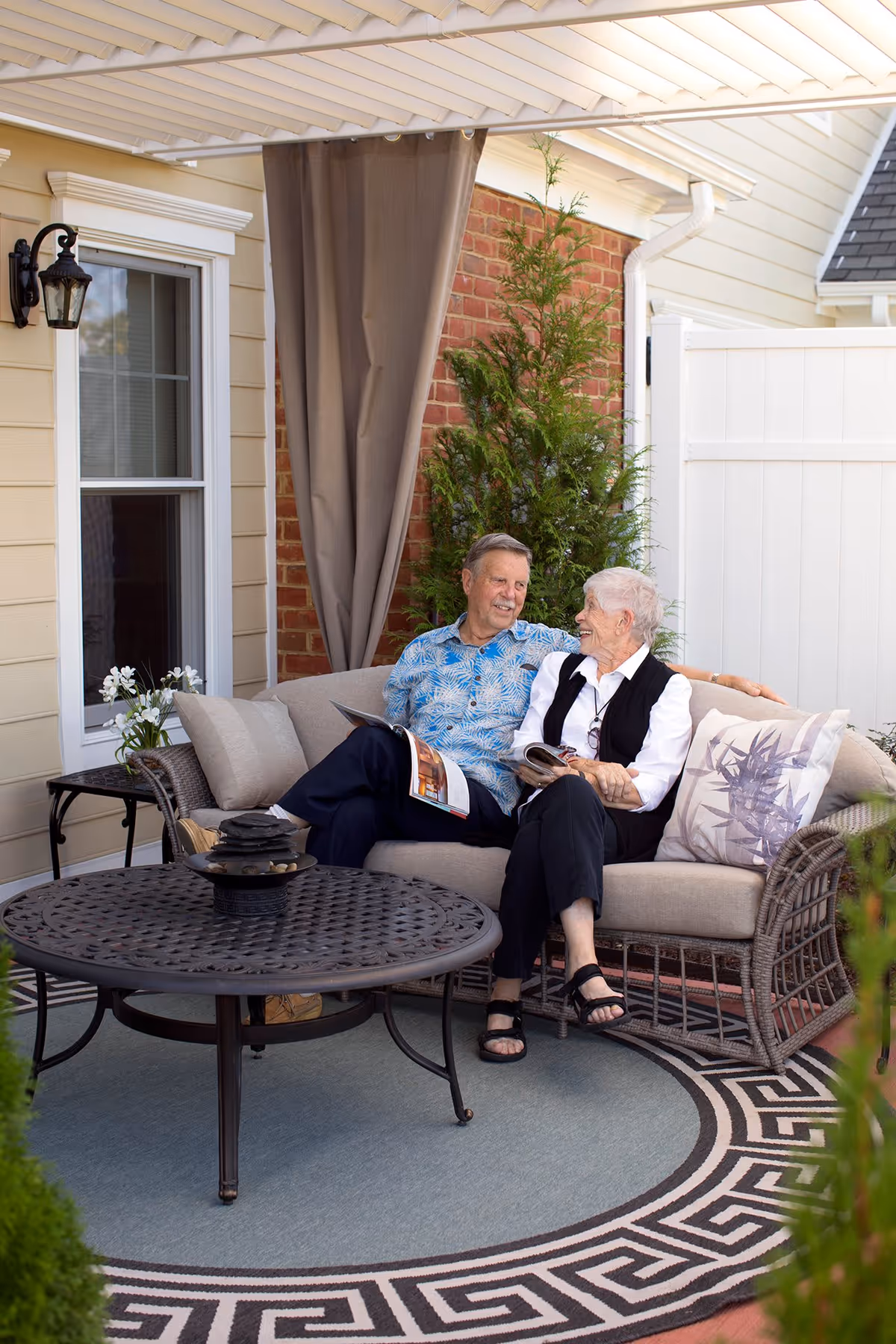 An elderly man and woman sitting together on a cushioned outdoor wicker sofa under a covered patio. They are smiling and holding magazines, surrounded by a round metal table, potted plants, and a decorative outdoor rug.