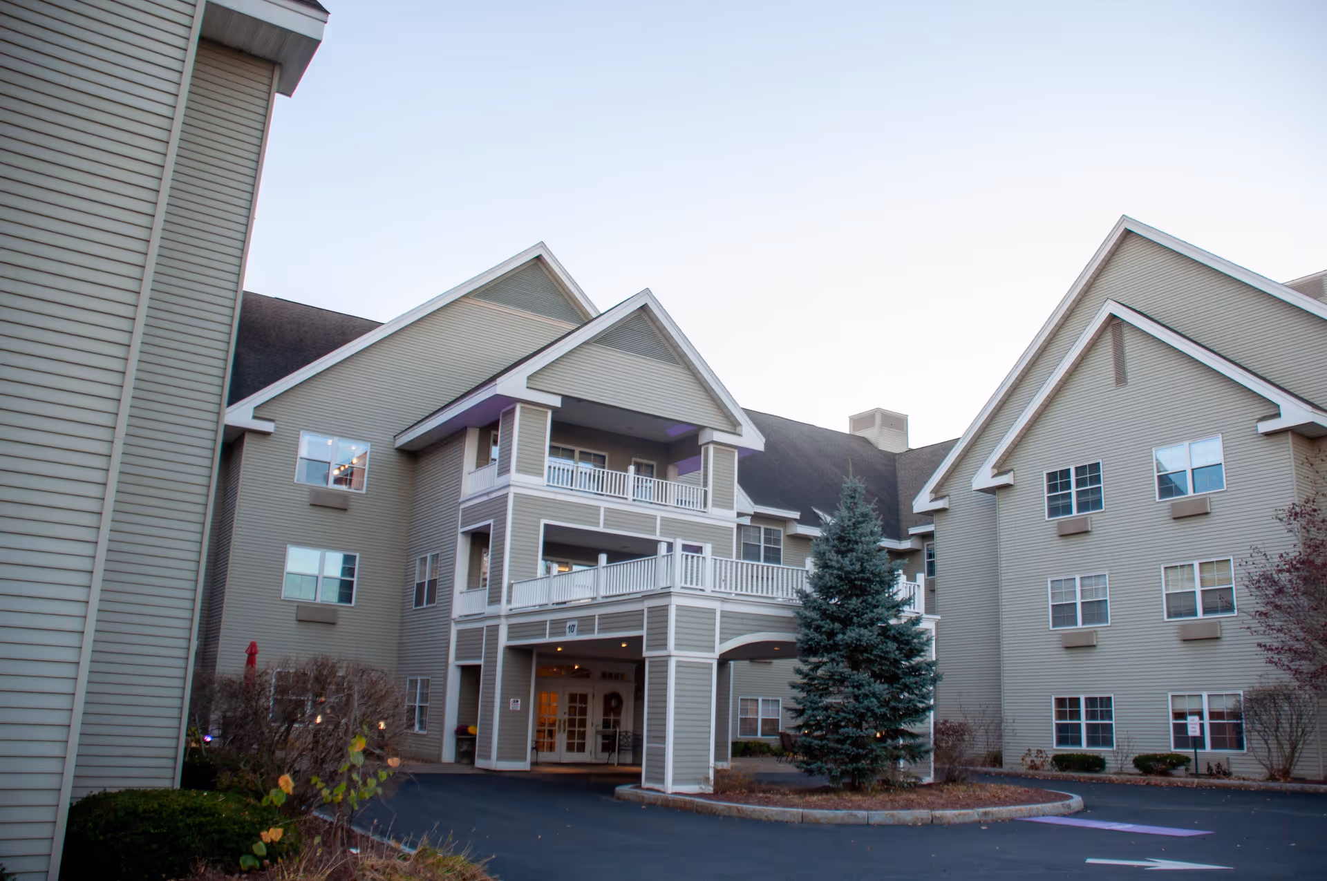 Exterior view of a multi-story senior living facility building with beige siding, multiple windows, and a covered entrance with balconies above. There is a small landscaped area with a tree and shrubs in front of the entrance.