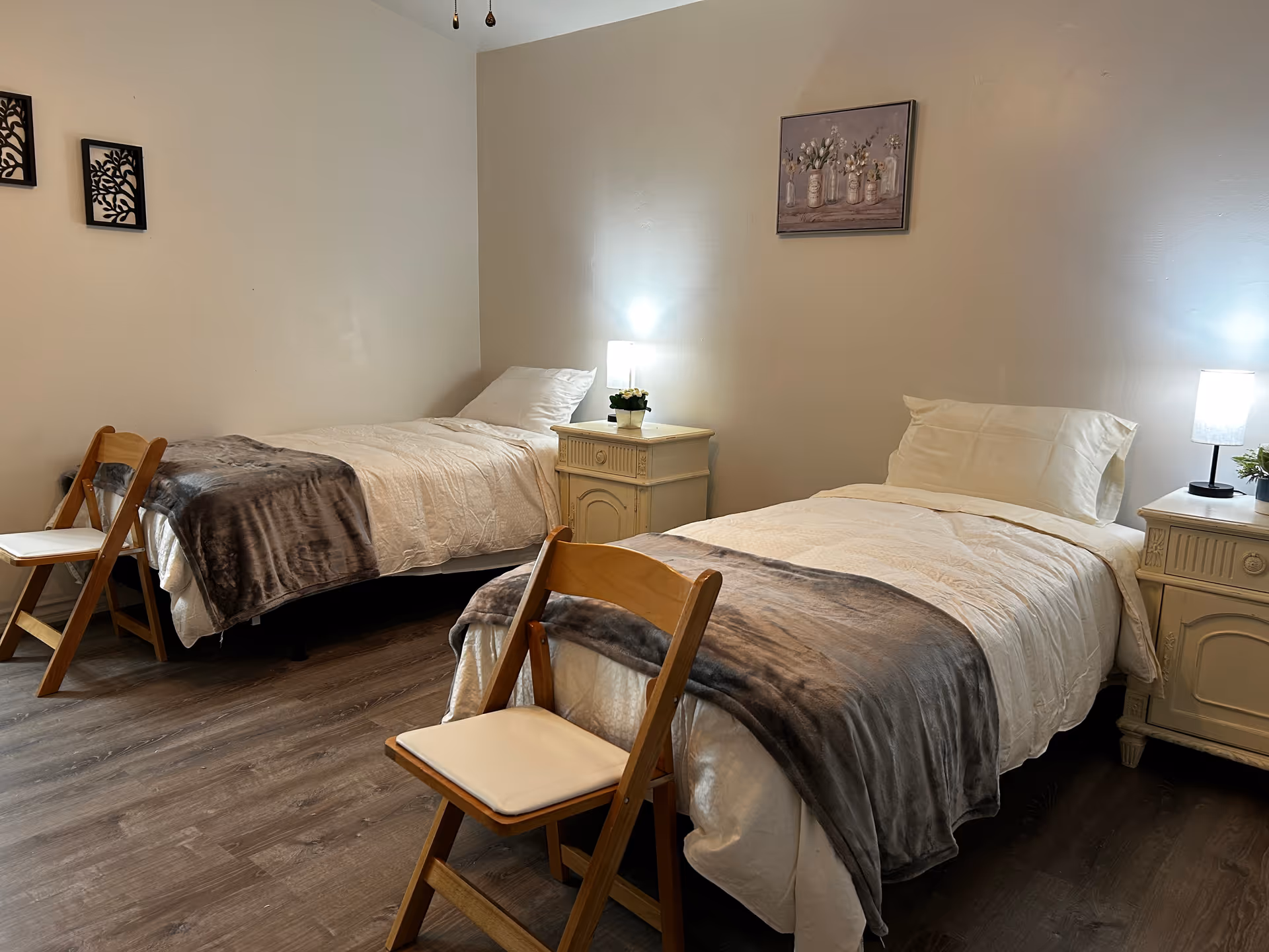A cozy bedroom with two single beds, each with white bedding and gray blankets. Each bed has a wooden chair placed at the foot. There are two matching nightstands between and beside the beds, each with a small lamp and a potted plant. The walls are light-colored with simple framed artwork hanging above the beds. The floor is a wood laminate.