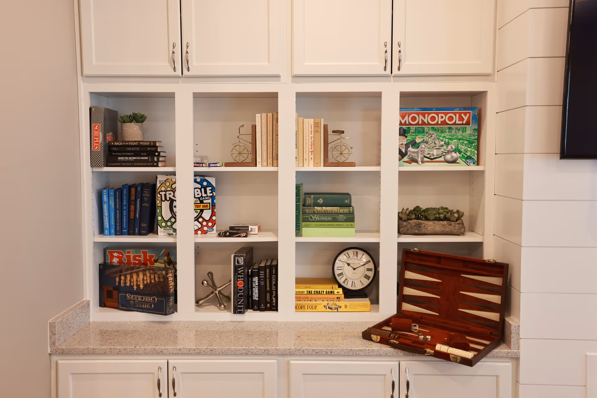 White built-in shelves filled with books, board games including Monopoly, Trouble, Risk, and Chess & Checkers, decorative items like a clock, small plants, and a backgammon set on the countertop.