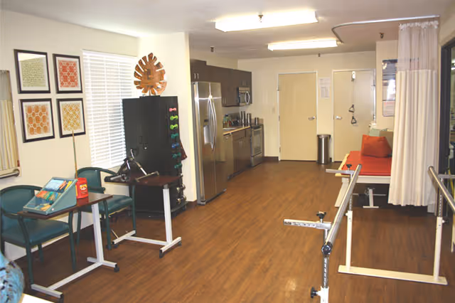A rehabilitation room with wooden flooring featuring parallel bars for walking exercises, a red cushioned therapy table with a privacy curtain, a kitchenette area with stainless steel appliances including a refrigerator, microwave, and dishwasher, and seating with green chairs and small tables. The walls have framed artwork and a rack with colorful dumbbells.