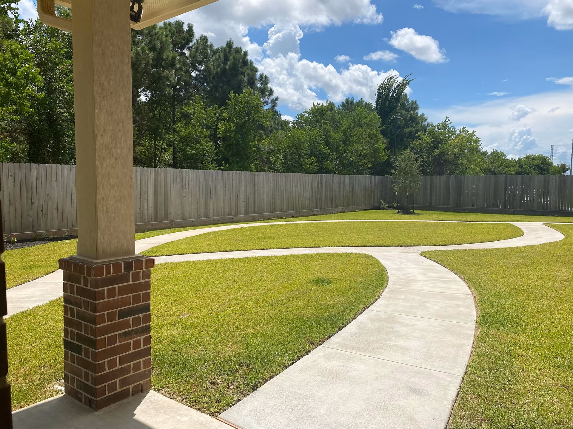 A sunny outdoor garden area with a curved concrete walkway surrounded by green grass. There is a wooden fence enclosing the space and trees in the background under a partly cloudy blue sky. A brick and beige column is visible in the foreground.