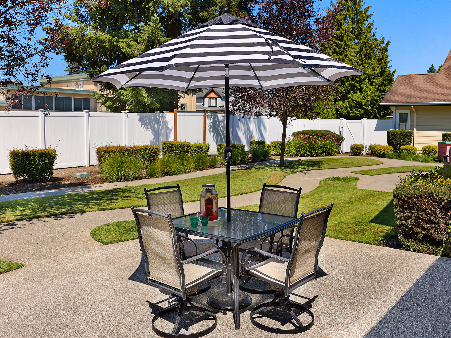 Outdoor patio with a glass-top table, four chairs and a black-and-white striped umbrella on a paved area surrounded by lawn, shrubs, and a white fence.