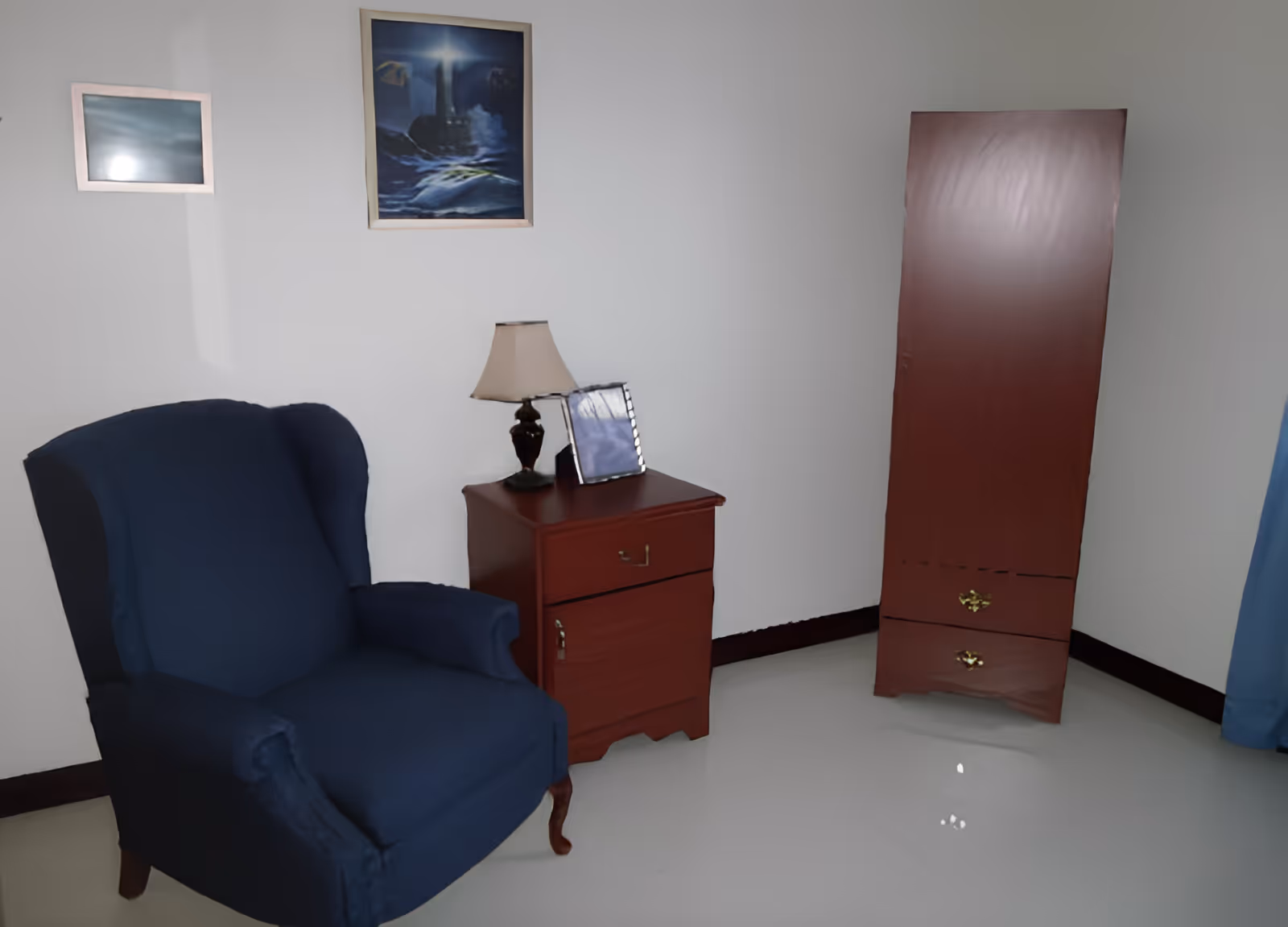 A cozy corner of a room featuring a dark blue upholstered armchair, a wooden nightstand with a table lamp and a framed photo, and a tall wooden cabinet with two drawers. Two framed pictures hang on the white wall above the furniture, and a blue curtain is partially visible on the right side.