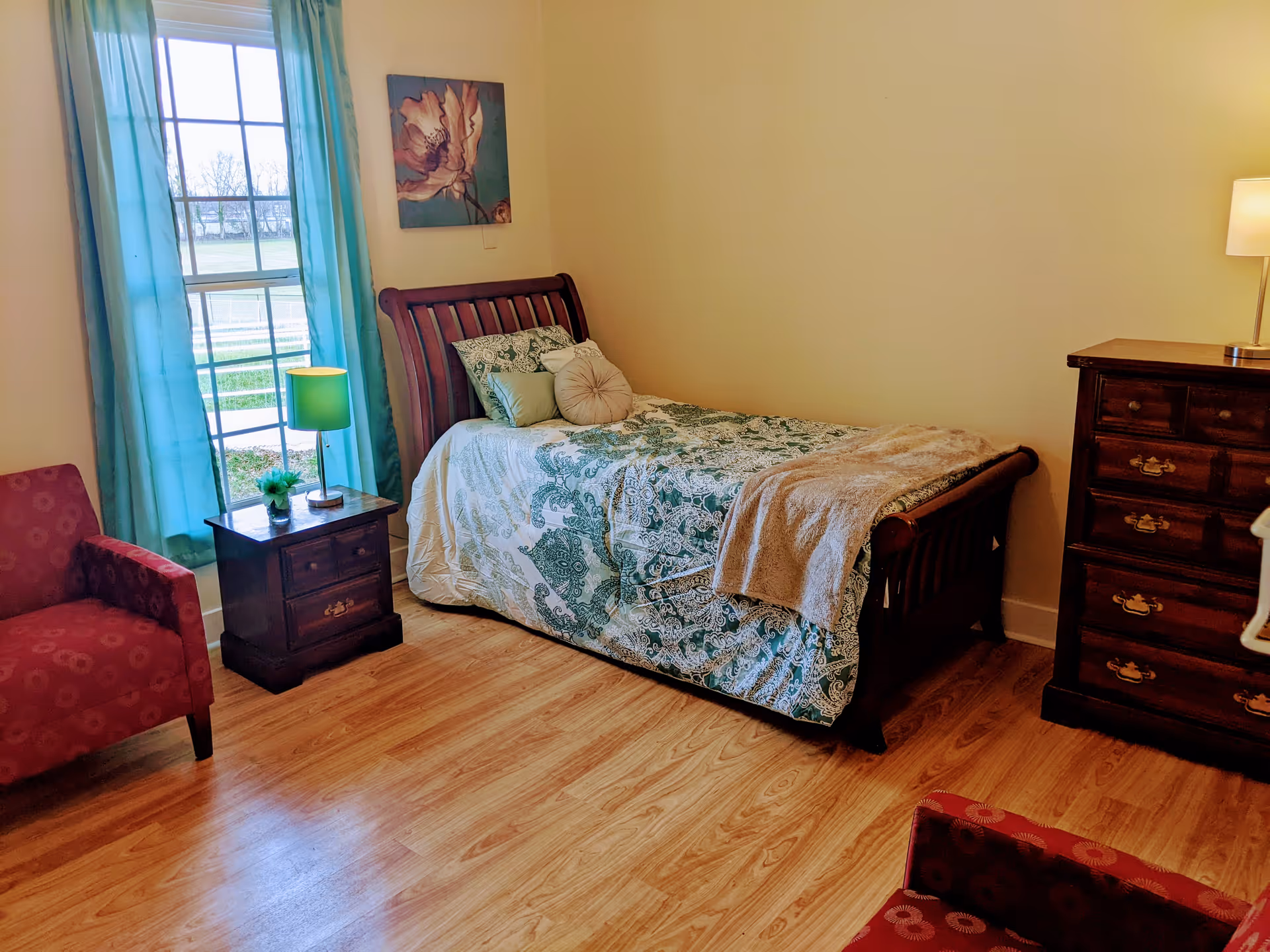 Sunny bedroom with a twin bed dressed in patterned bedding, wooden nightstand and dresser, teal curtains, and two red armchairs.