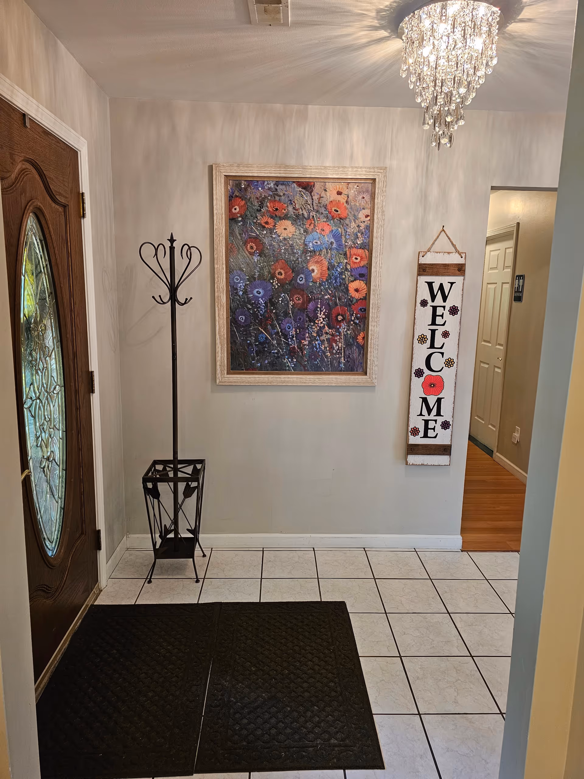 Entryway of Orchard Meadows Senior Care Home featuring a wooden door with an oval glass panel, a black metal coat rack with an umbrella stand, a colorful floral painting on the wall, a vertical welcome sign with flowers, white tiled floor with two black mats, and a crystal chandelier ceiling light.