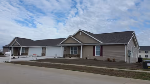 Single-story residential building with beige siding and a dark shingled roof under a partly cloudy sky. The building has a garage and a small covered porch area. The surrounding area includes a paved driveway and bare soil landscaping.