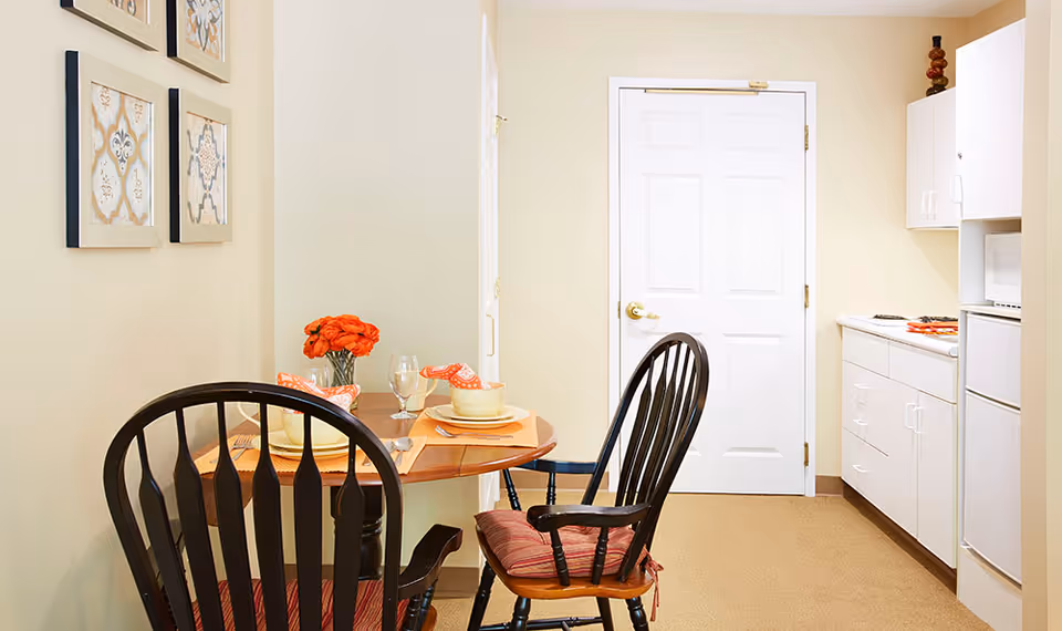 Small dining nook with a round table set for two and wooden chairs beside a compact kitchenette and a white door.