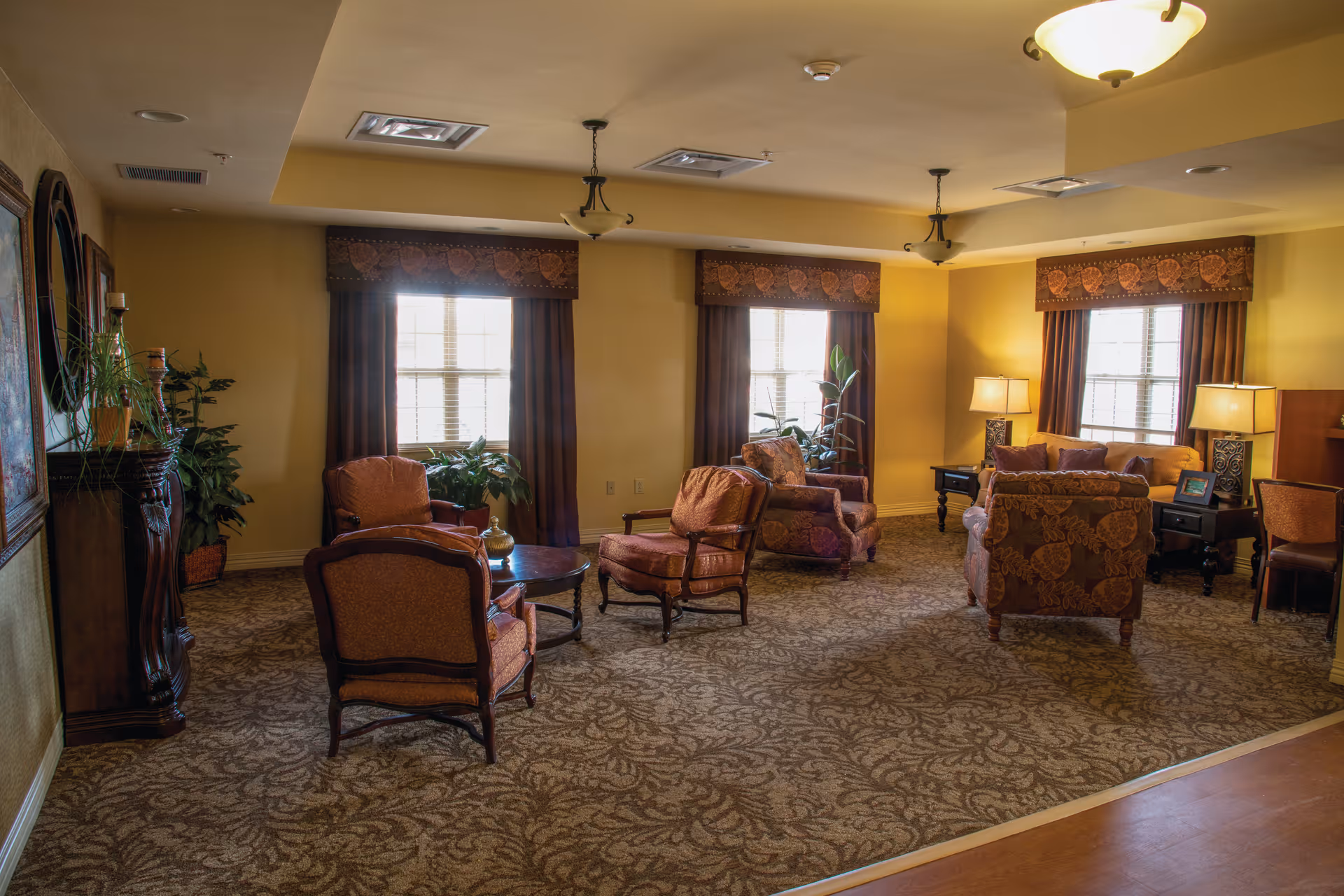 A cozy living room area in Cabell Healthcare Center with multiple upholstered armchairs and sofas arranged around small tables. The room features patterned carpet, yellow walls, three windows with dark curtains and valances, potted plants, table lamps, and ceiling light fixtures providing warm lighting.