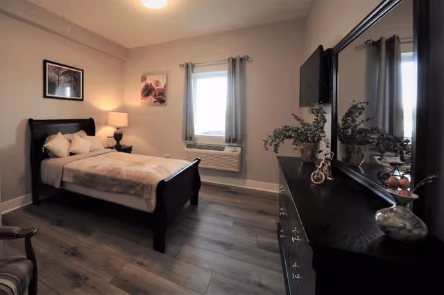 A cozy bedroom with a dark wooden bed frame, a bed with beige bedding and pillows, a nightstand with a lamp, two framed pictures on the wall, a window with gray curtains, and a dark wooden dresser with a large mirror and decorative items including plants and a small bicycle figurine.
