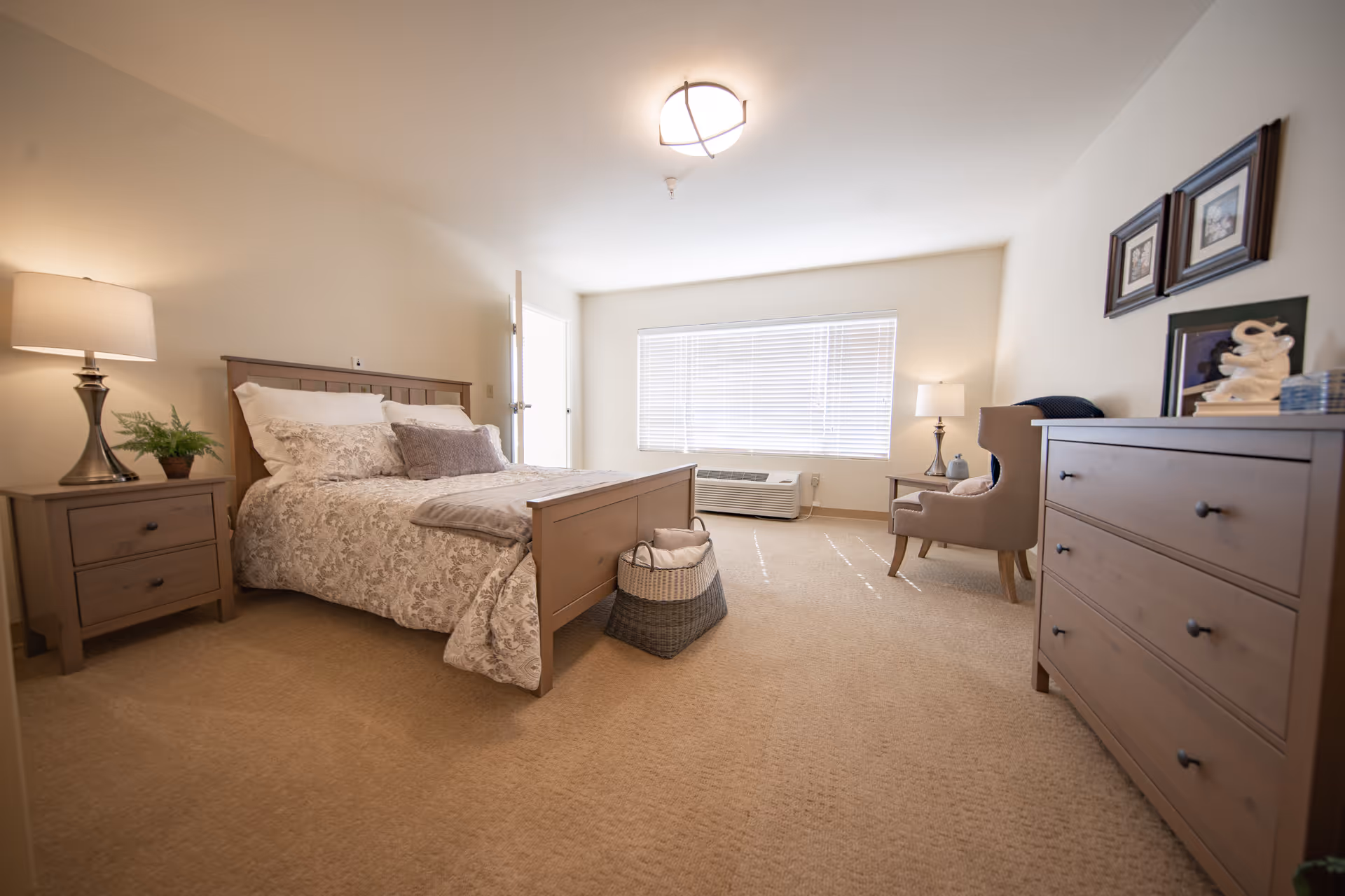 A well-lit bedroom with a wooden bed frame, floral bedding, and multiple pillows. There is a wooden nightstand with a lamp and a small plant on the left side of the bed. A basket is placed at the foot of the bed. On the right side of the room, there is a wooden dresser with decorative items and framed pictures above it. A comfortable armchair with a side table and lamp is positioned near a large window with blinds. The room has beige carpeting and neutral-colored walls.
