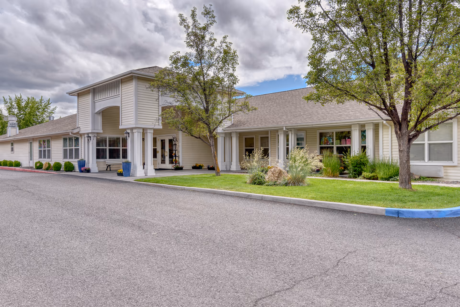 Exterior front entrance of a single-story assisted living facility with a covered portico, lawn, and trees.
