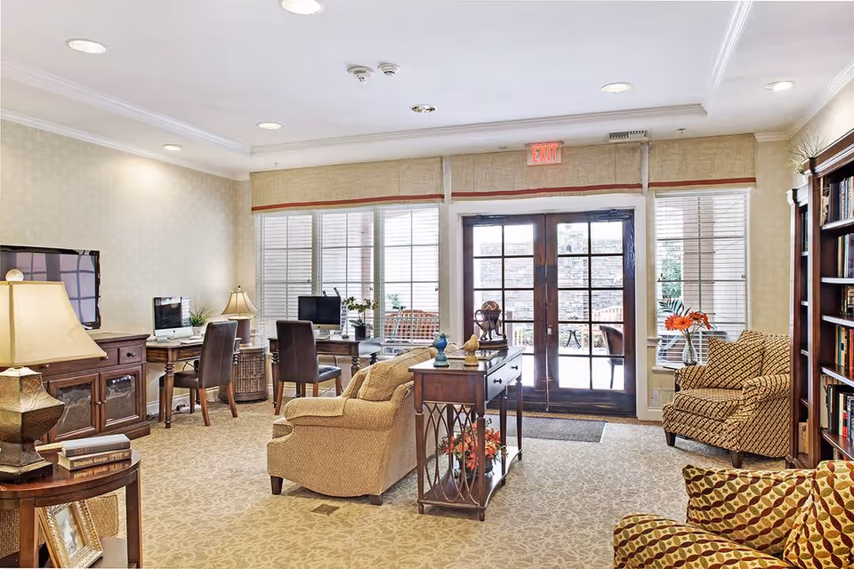 A bright communal living room with armchairs, sofas, desks, bookshelves and French doors leading outside.