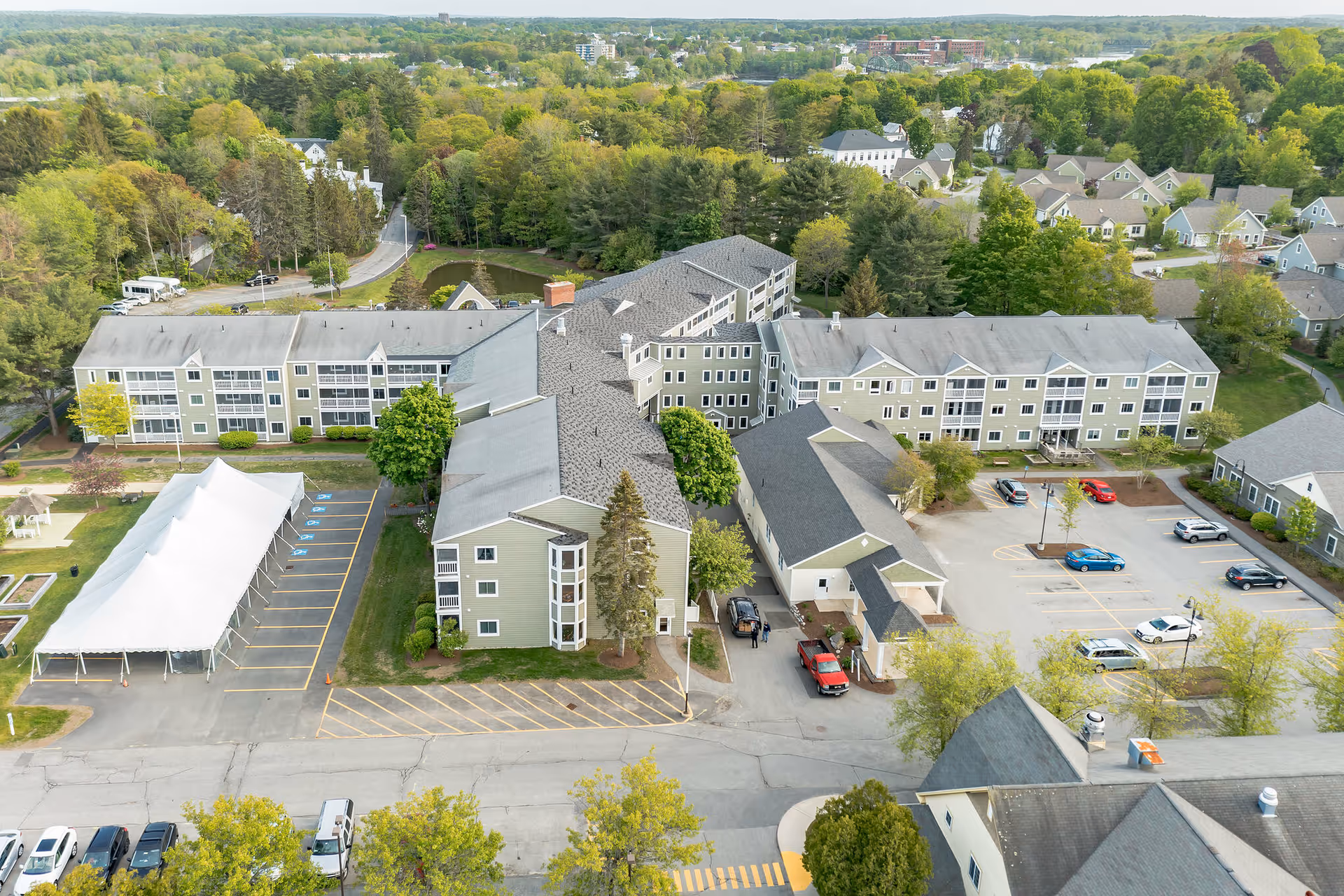 Aerial view of The Highlands senior living facility showing multiple connected buildings with gray roofs, surrounded by trees and greenery. There is a parking lot with several cars, a large white event tent, and residential houses in the background.