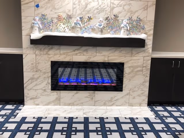 A modern electric fireplace set in a marble-tiled wall with a black mantel shelf above it. On the mantel are decorative snowmen and colorful snowflake ornaments arranged on a layer of artificial snow. The floor in front of the fireplace has a blue and white patterned carpet, and there are dark cabinets on either side of the fireplace.