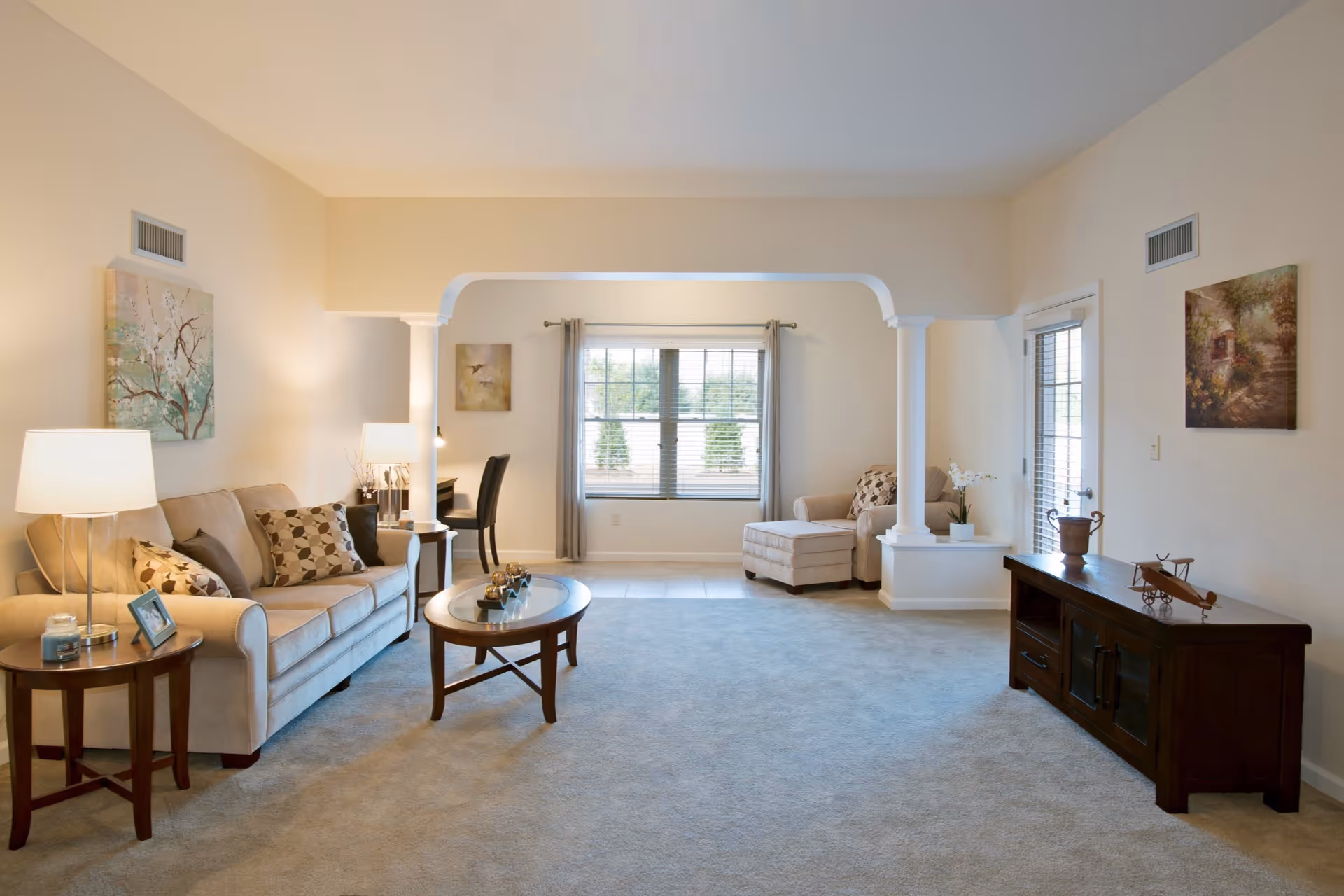 A spacious living room with beige walls and carpet. The room features a beige sofa with patterned cushions, a wooden coffee table, a side table with a lamp and framed photo, and a dark wooden TV stand with decorative items. There is a large window with curtains, a comfortable armchair with an ottoman, and a small desk with a chair in the corner. The room is well-lit with natural light and has two white columns framing the window area.
