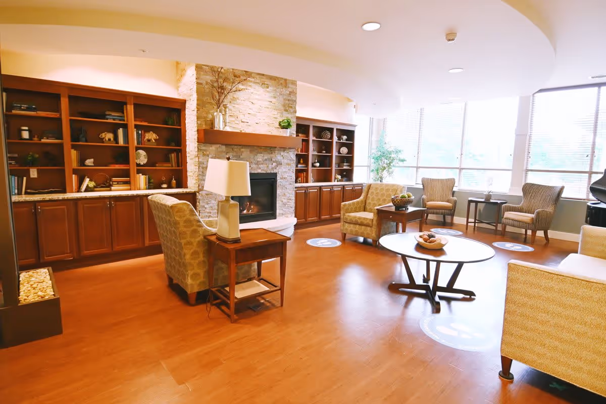A cozy living room area in a senior living facility with wooden flooring, a stone fireplace, built-in wooden bookshelves, several upholstered armchairs, a round coffee table with a decorative bowl, and large windows letting in natural light.