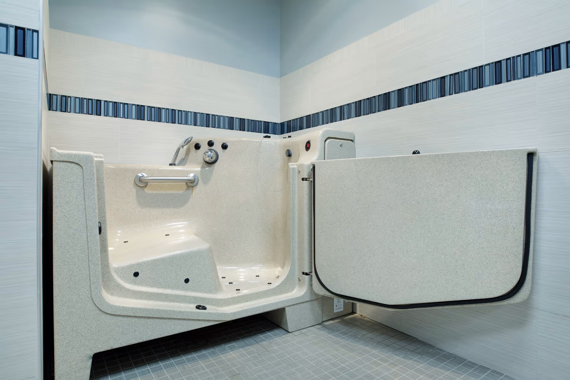 A walk-in bathtub with an open door designed for easy access, featuring built-in seat, grab bar, and control knobs, set against tiled walls with a decorative horizontal stripe.