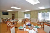Bright dining room with multiple round tables set with plates and napkins, wooden chairs, windows with valances, a wall-mounted TV and a grandfather clock.