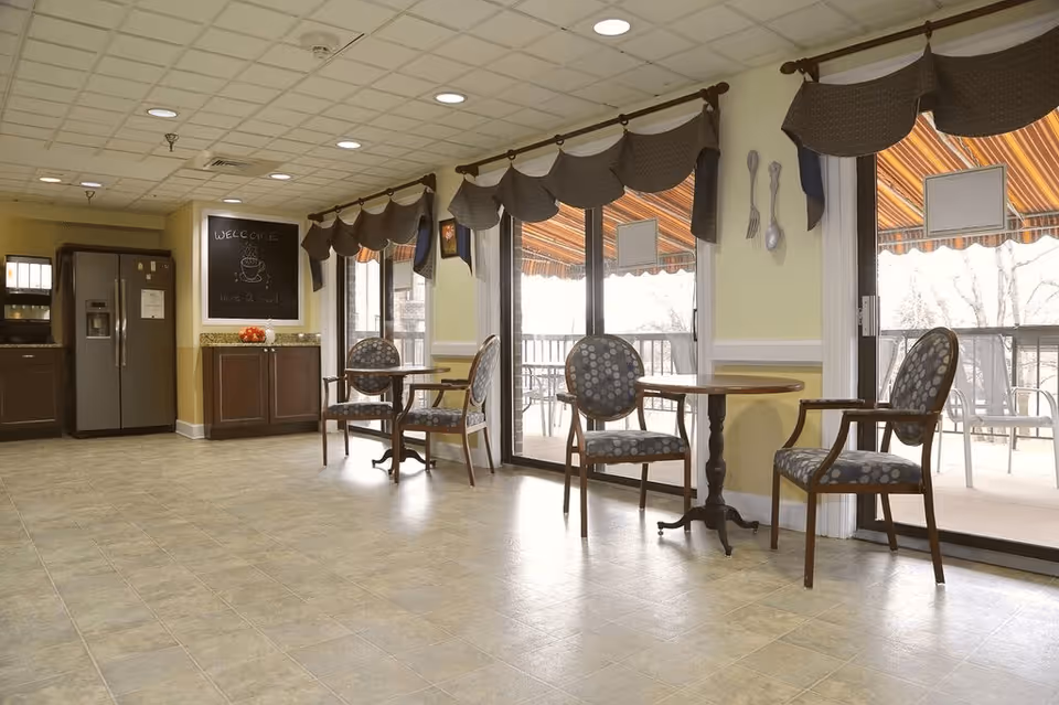 A bright dining area with tiled floors, featuring round tables each paired with two patterned chairs. Large sliding glass doors with brown valances lead to an outdoor patio with chairs. A kitchen area with a refrigerator and cabinets is visible in the background, along with a chalkboard sign that says 'WELCOME'.