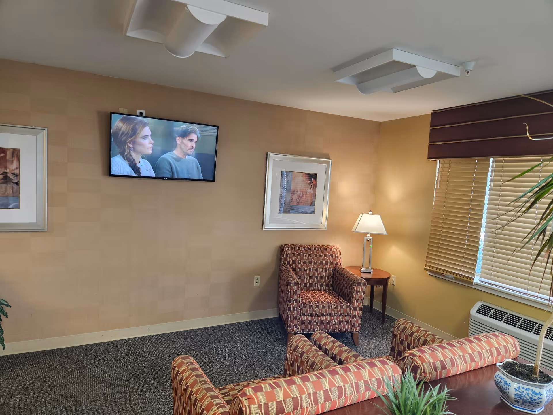 Small sitting area with patterned armchairs, a wall-mounted TV, side table with lamp, and window blinds.