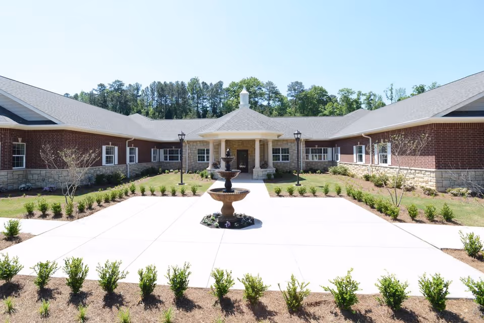 Front exterior view of a single-story senior living facility building with a central entrance featuring a covered porch supported by columns, a tiered water fountain in the foreground, neatly trimmed bushes, and a clear blue sky with trees in the background.
