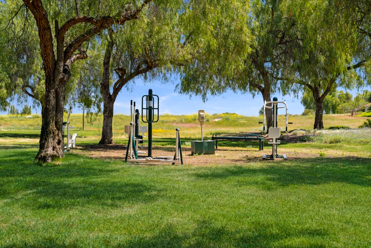Outdoor exercise equipment set up on a grassy area under large trees with a clear blue sky in the background at Westmont of Riverside.