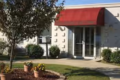 Entrance to a building with a red awning above glass double doors. The awning has text that is partially visible. There are bushes and a tree near the entrance, along with flower pots on a mulched area beside a concrete walkway.