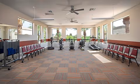 A spacious exercise room with large windows letting in natural light. The room contains several pieces of cardio equipment including treadmills, stationary bikes, and elliptical machines. Along the walls, there are rows of red chairs and a rack holding blue exercise balls. Ceiling fans and overhead lights are visible, and outside the windows, palm trees and greenery can be seen.