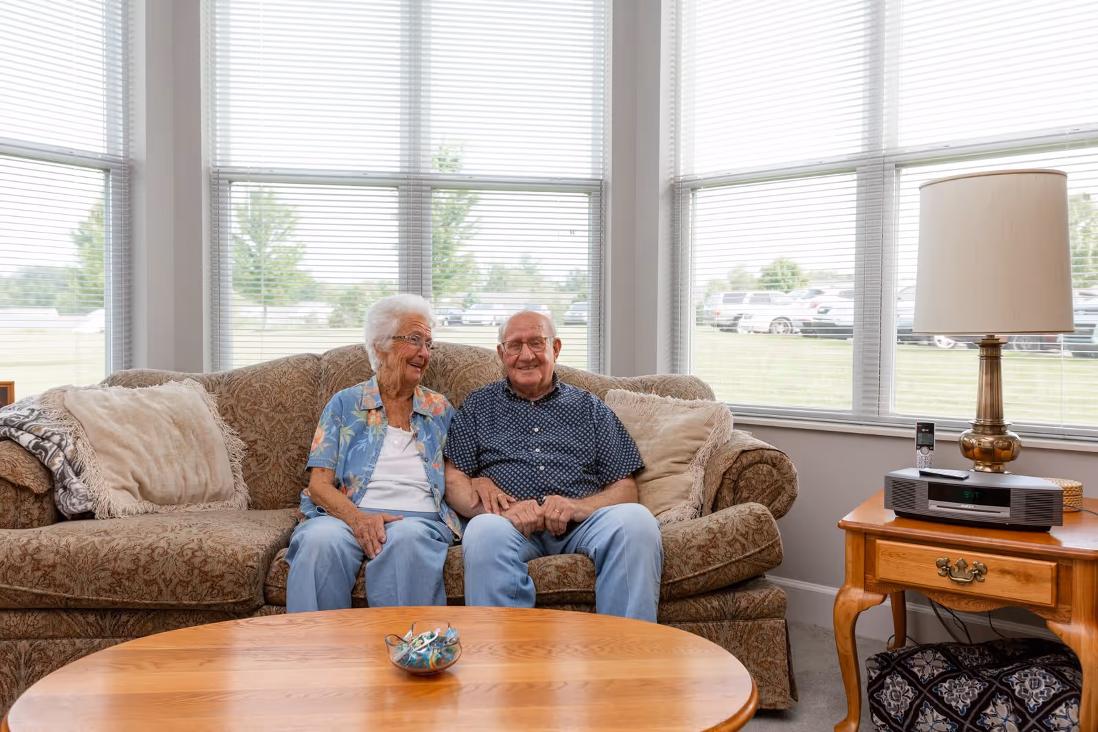 An elderly couple sitting closely together on a patterned sofa in a bright room with large windows behind them. The woman is wearing a blue floral shirt and light blue pants, while the man is wearing a dark blue patterned shirt and light blue pants. There are beige pillows on the sofa, a wooden coffee table with a small bowl of candy in front of them, and a wooden side table with a lamp and a stereo system to the right.