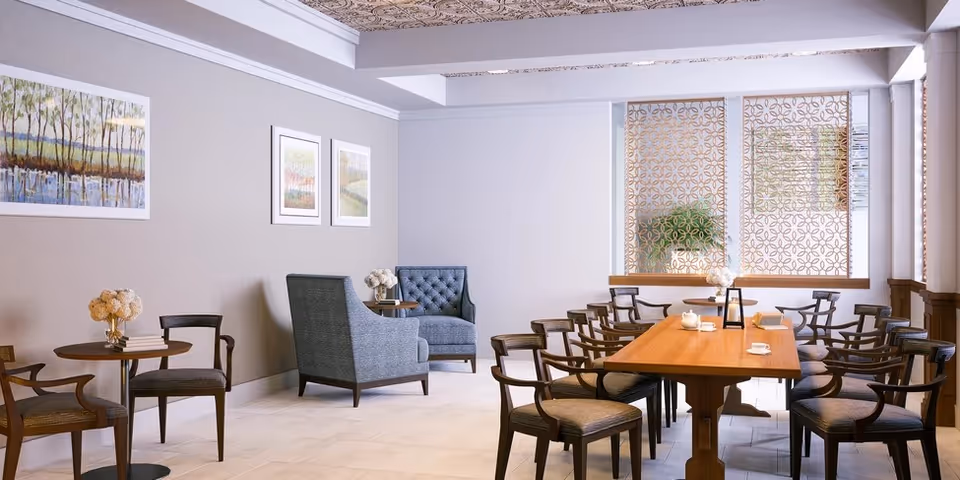 A well-lit common area with a long wooden table surrounded by multiple wooden chairs with cushions. To the left, there are two small round tables each with two chairs and a vase of flowers. The walls are decorated with framed landscape paintings, and there is a patterned screen on the window letting in natural light.