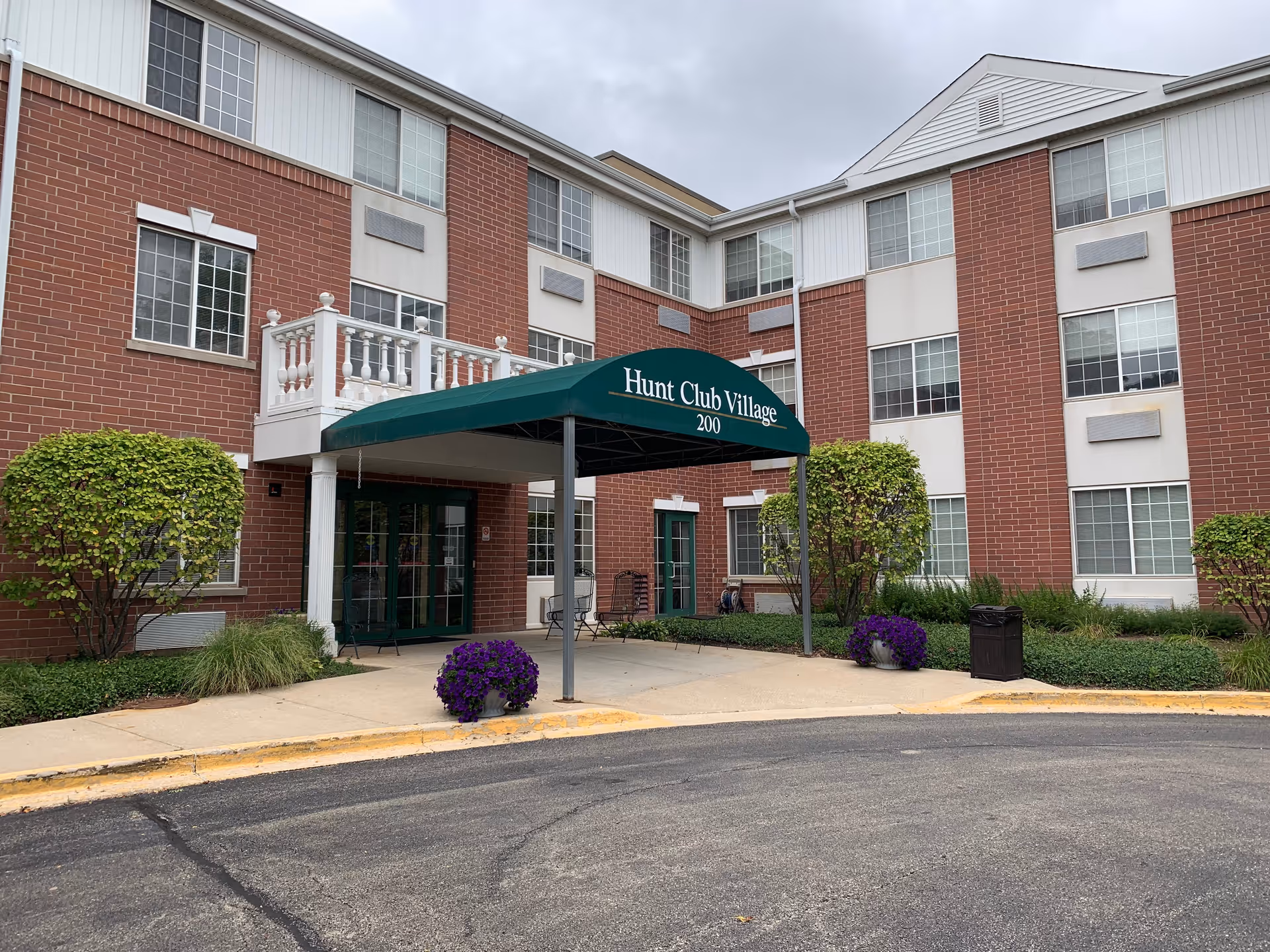 Front entrance of a brick senior living building with a green awning reading 'Hunt Club Village' and potted flowers by the walkway.