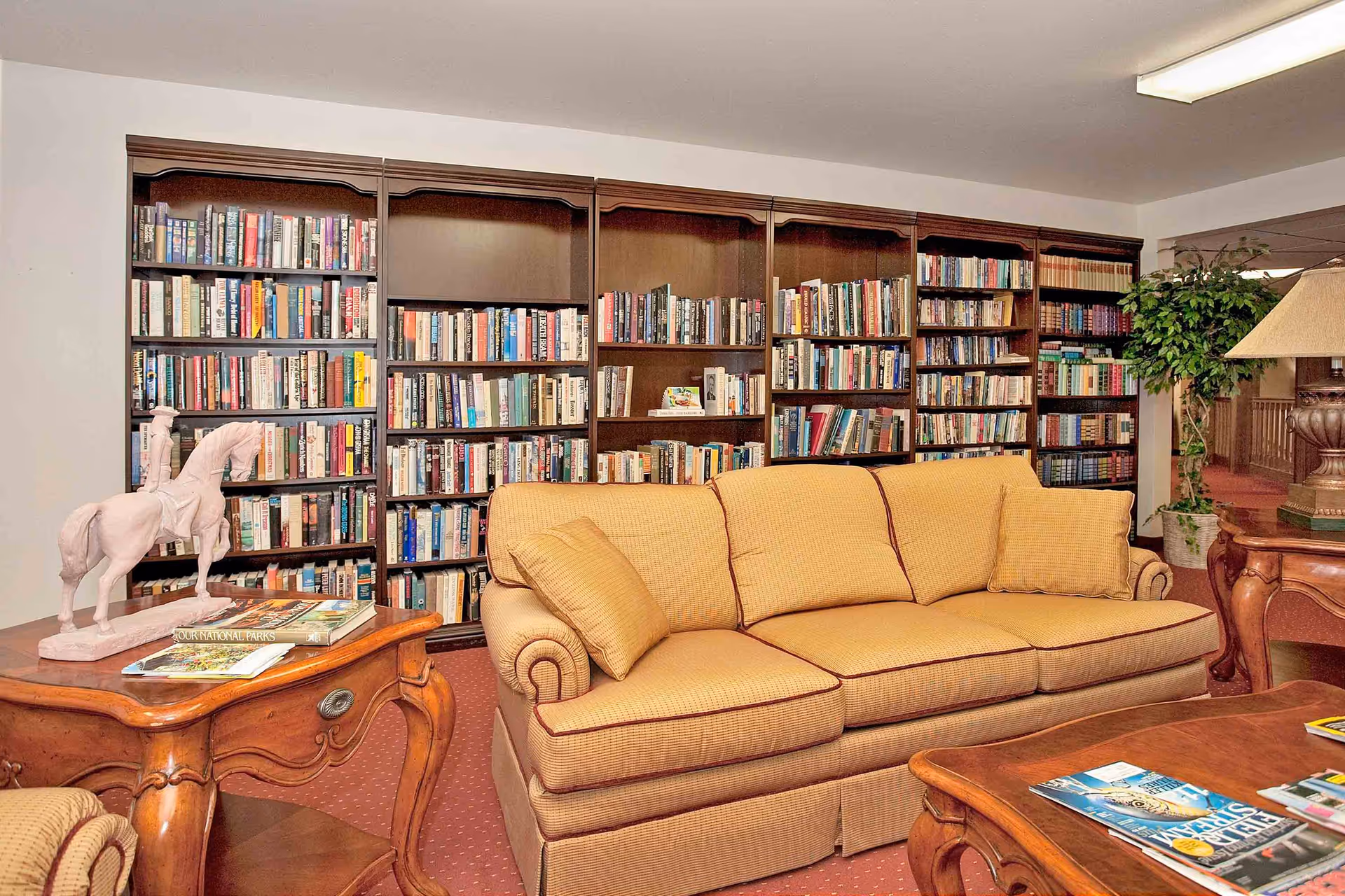 A cozy living room area in a senior living facility featuring a large beige sofa with matching cushions, wooden coffee and side tables with magazines and a white horse statue, a tall bookshelf filled with books, a potted plant, and a lamp with a beige shade.
