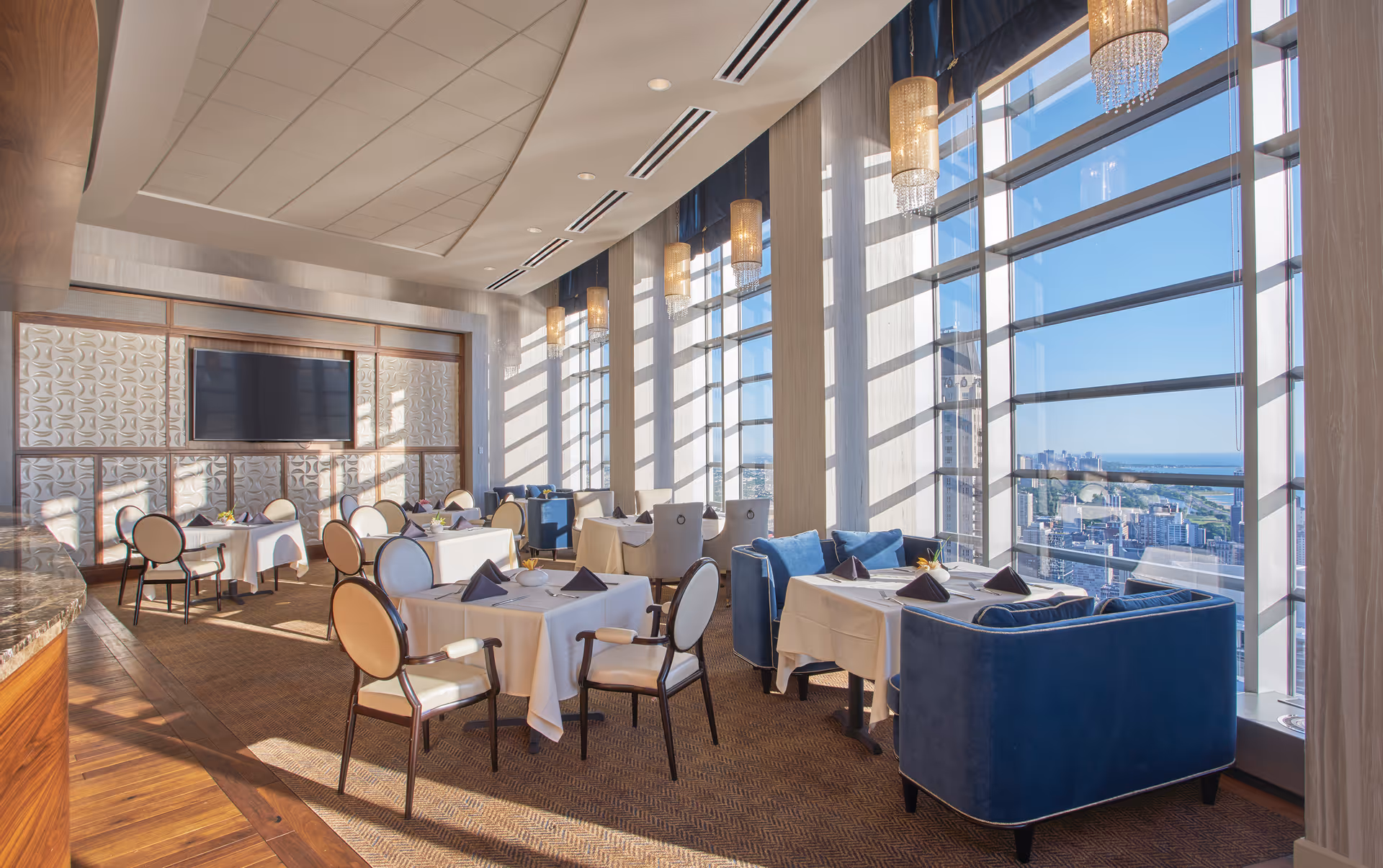 Sunlit upscale dining room with tables set, blue armchairs and floor-to-ceiling windows overlooking the city.