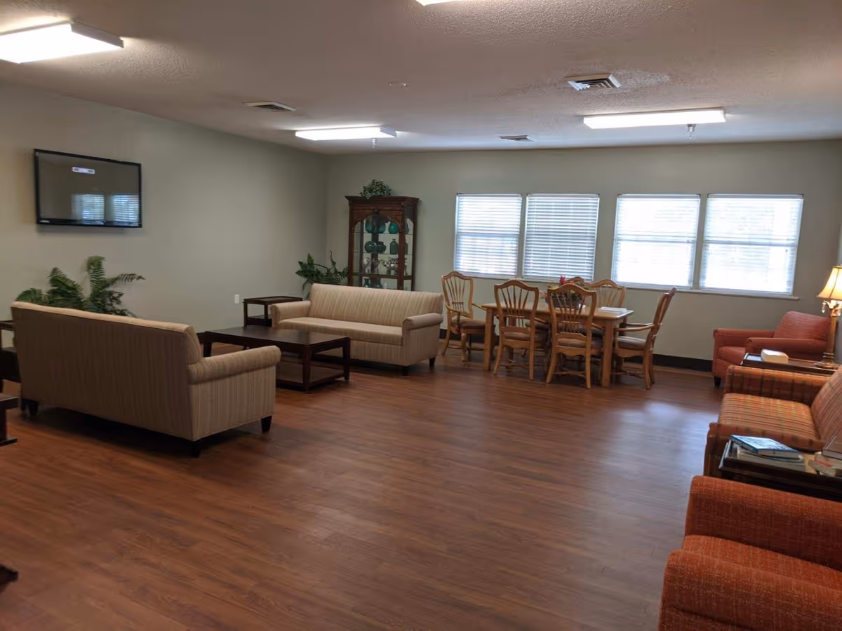 A spacious living room area with wooden flooring, two beige sofas facing each other with a dark wooden coffee table in between, a wooden dining table with six chairs near three windows with blinds, a wooden display cabinet with decorative items, and two orange armchairs with side tables and a lamp.