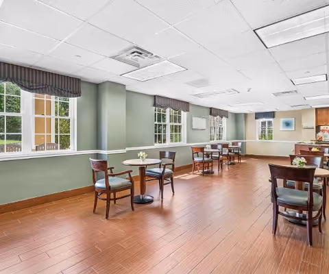 A bright dining room with several small round tables, each surrounded by wooden chairs with green cushions. The room has large windows with striped valances, light green walls with white trim, and a wooden floor. There are small flower arrangements on the tables and framed artwork on the walls.