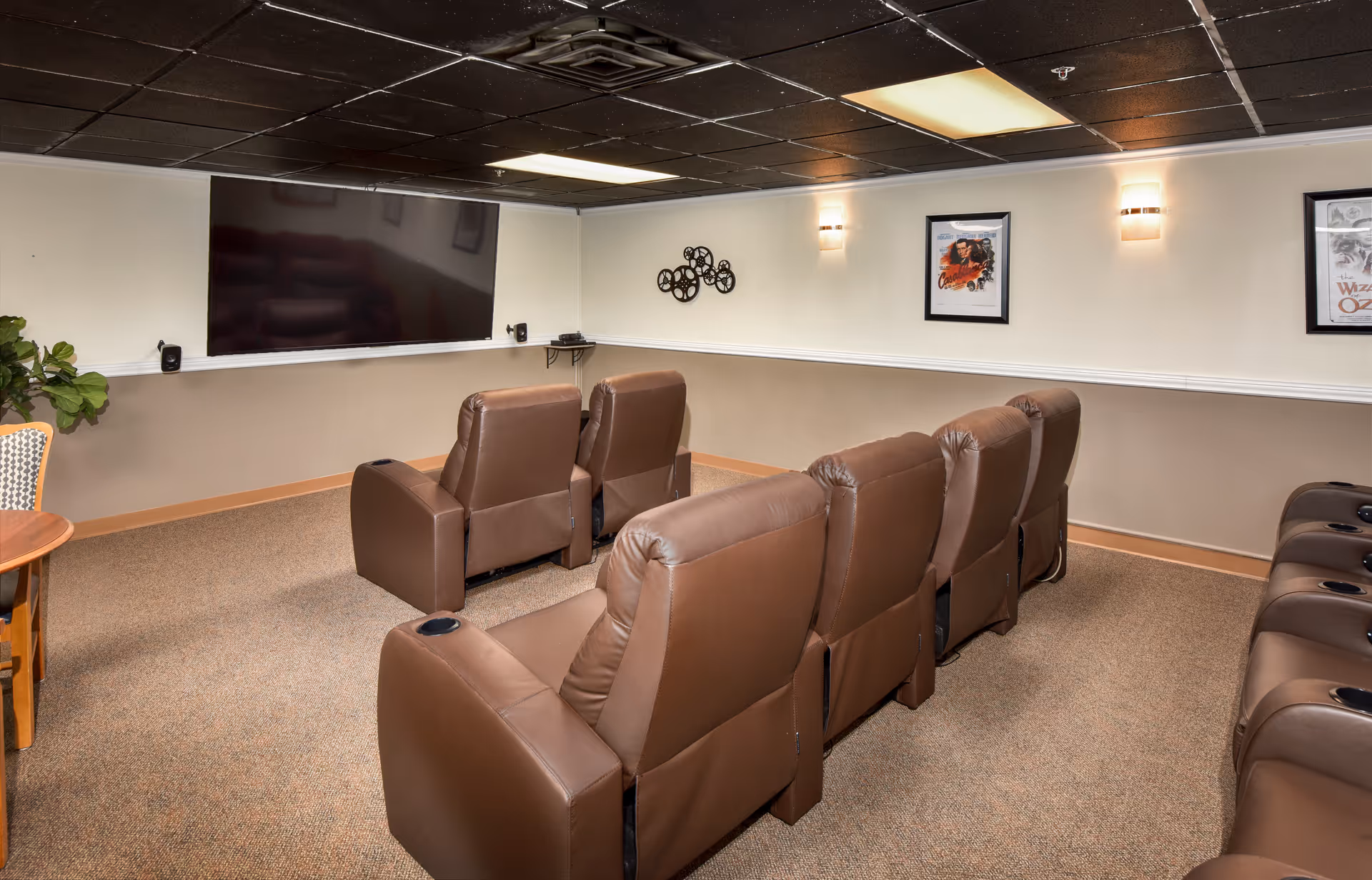 A cozy media room with two rows of brown leather recliners facing a large flat-screen TV mounted on the wall. The room has beige walls with a white chair rail, carpeted floor, and a black ceiling with recessed lighting. There are framed movie posters on the walls and a small round wooden table with a patterned chair in the corner.