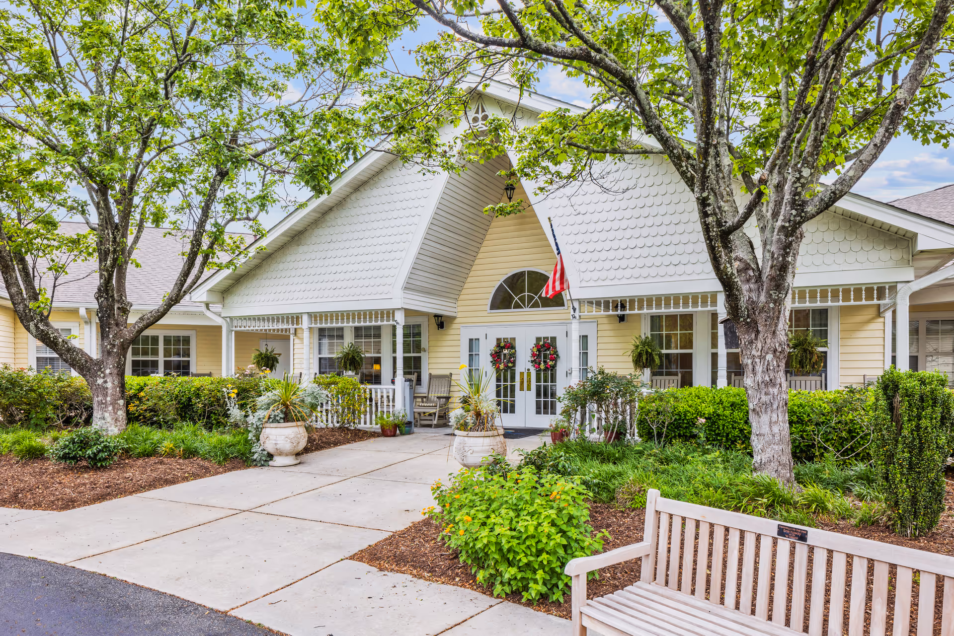 Front exterior view of TerraBella Southport facility showing a yellow building with white trim, a peaked roof, and double glass doors decorated with wreaths. There are two large trees, green shrubs, potted plants, and a wooden bench in the landscaped area near the entrance. An American flag is displayed above the doors.