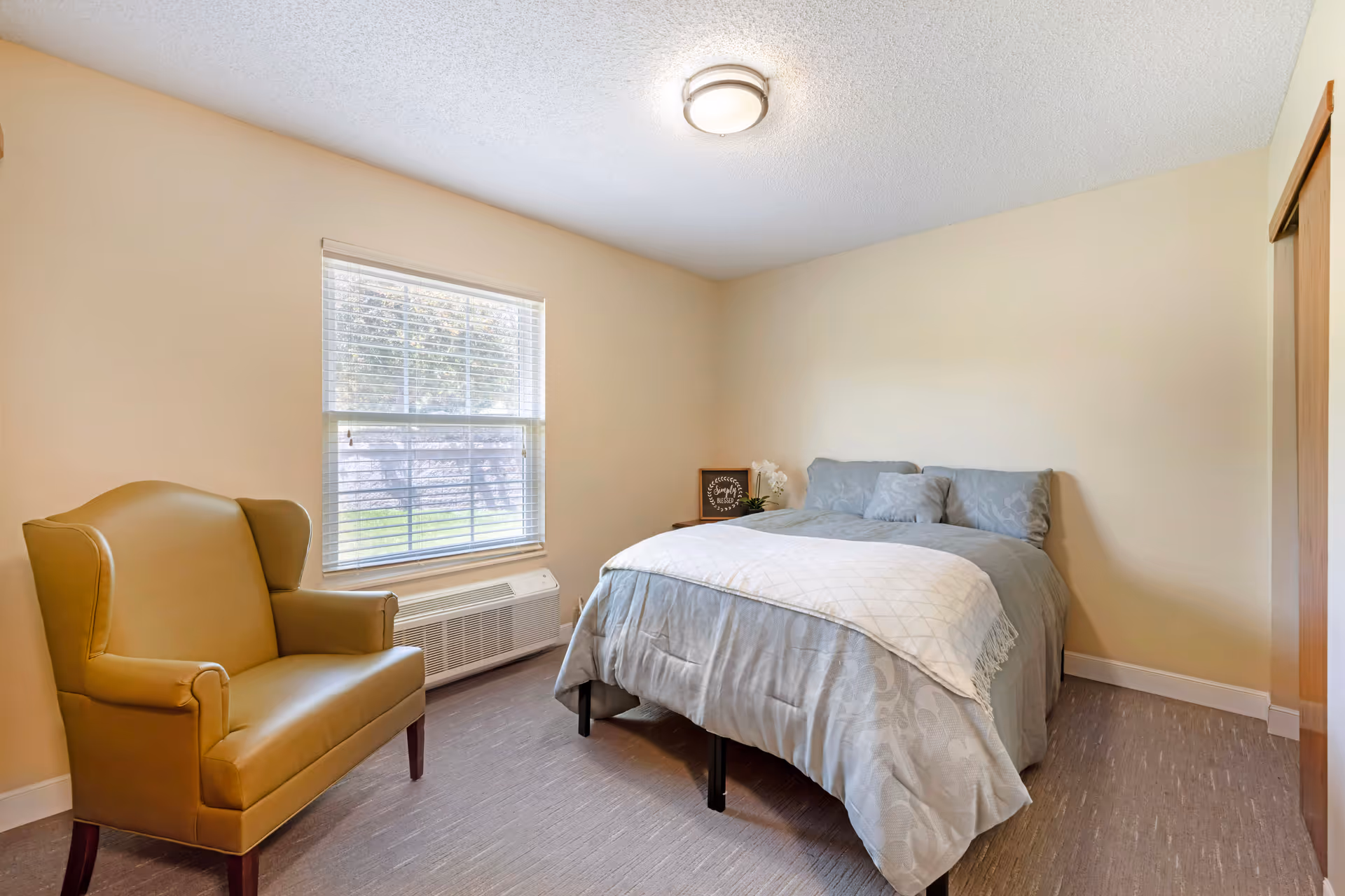 Small bedroom with a made bed, a yellow armchair beside a window, and a bedside table.