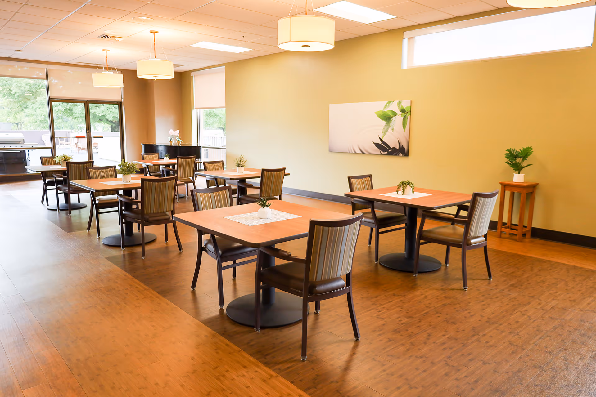 A bright dining room with several wooden tables and chairs arranged neatly. Each table has a small plant centerpiece. Large windows allow natural light to fill the room, and there are modern ceiling lights. A painting with green leaves hangs on the beige wall, and there is a small wooden stand with a plant in the corner.