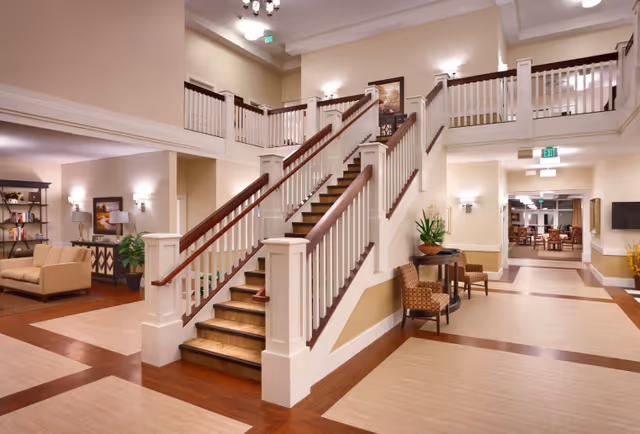 A spacious and well-lit interior of a senior living facility featuring a central staircase with wooden handrails and white balusters. The area includes seating with two patterned chairs and a small table with a plant, a cozy sitting area with a beige sofa and lamps, and a hallway leading to a dining area with tables and chairs. The walls are painted in neutral tones with warm lighting fixtures.