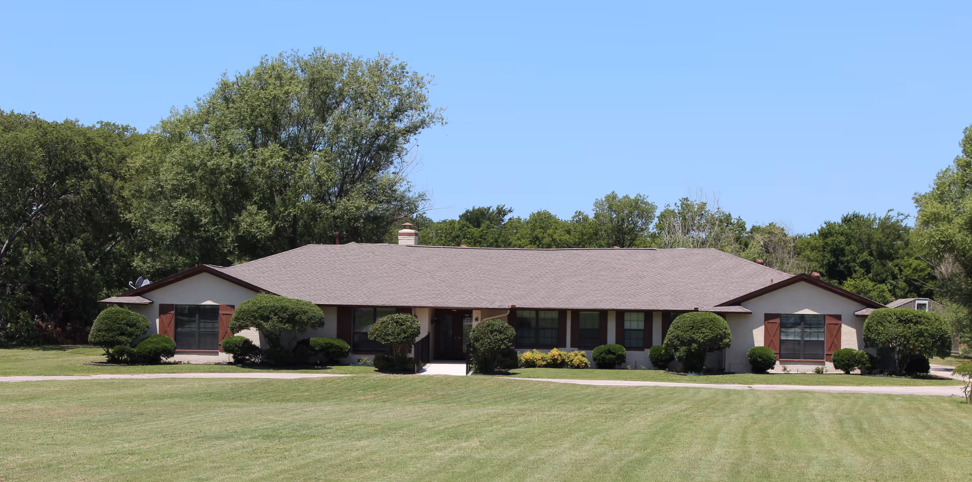 Single-story building with a brown roof and beige walls, surrounded by neatly trimmed bushes and trees, set against a clear blue sky with a large grassy lawn in the foreground.