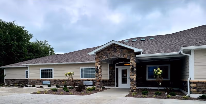 Exterior view of a single-story building with beige siding and stone accents around the entrance. The entrance has a covered porch with an archway supported by stone columns. There are several windows along the front of the building and small landscaped areas with shrubs and young trees. The sky is cloudy.