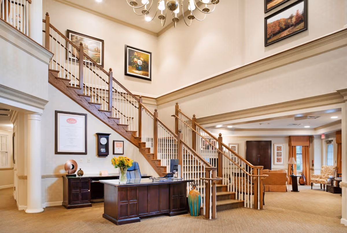 Bright lobby with a central reception desk and a grand wooden staircase leading to an upper level, with seating visible in the background.