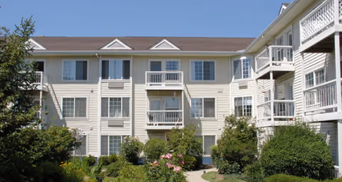 Exterior view of a multi-story senior living facility building with beige siding, white balconies, and multiple windows. The foreground features a landscaped garden with green bushes, trees, and flowering plants under a clear blue sky.