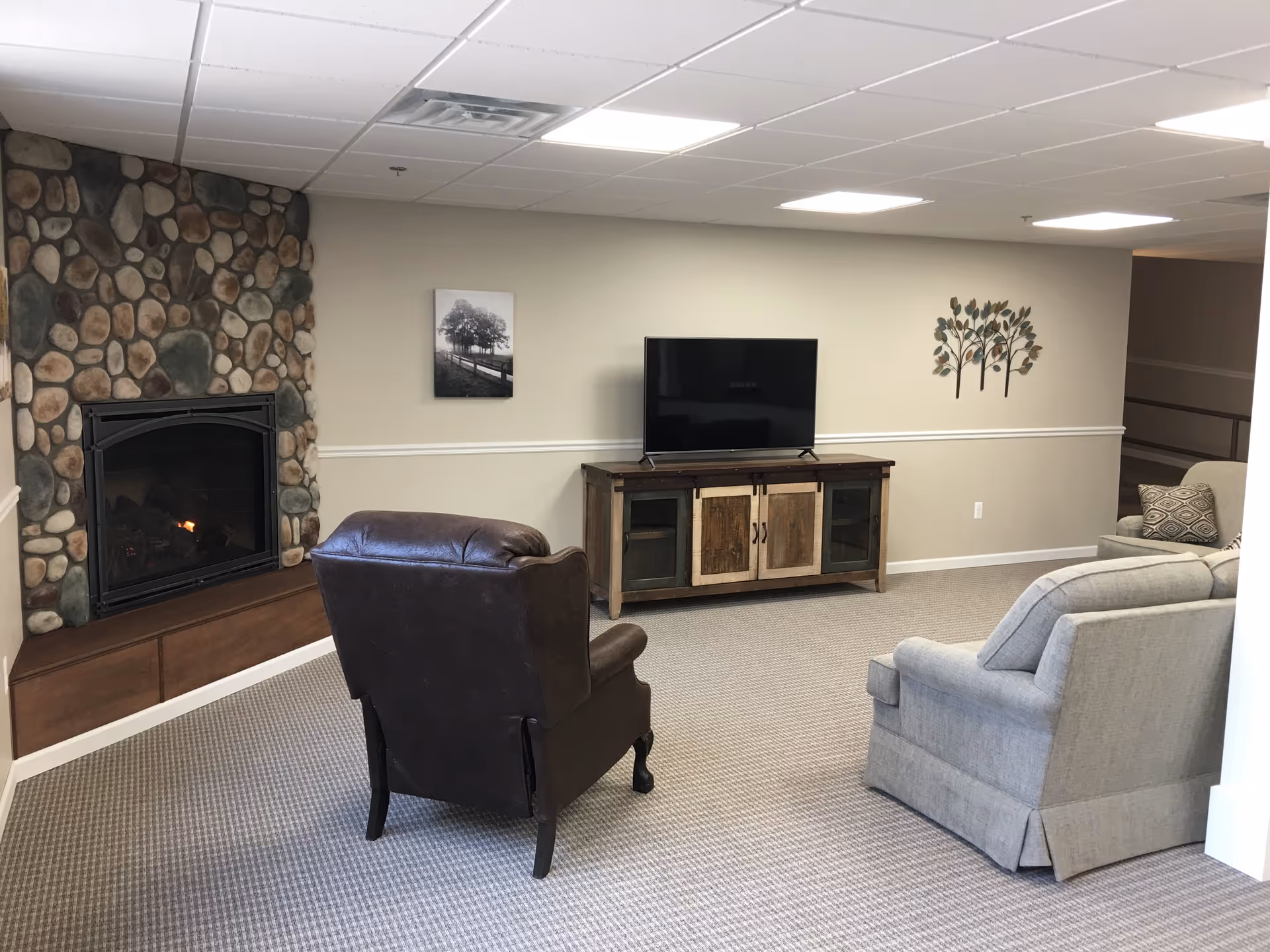 A cozy senior living room with a stone fireplace on the left, a brown leather armchair facing a flat-screen TV on a wooden cabinet, and two gray upholstered armchairs. The walls are beige with a white chair rail, decorated with a black and white photo and a metal wall art of trees.